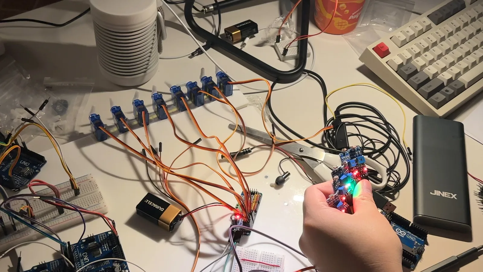 A desk at night. An Arduino Nano held in one hand is lit up, with a breadboard and six servos lined up.