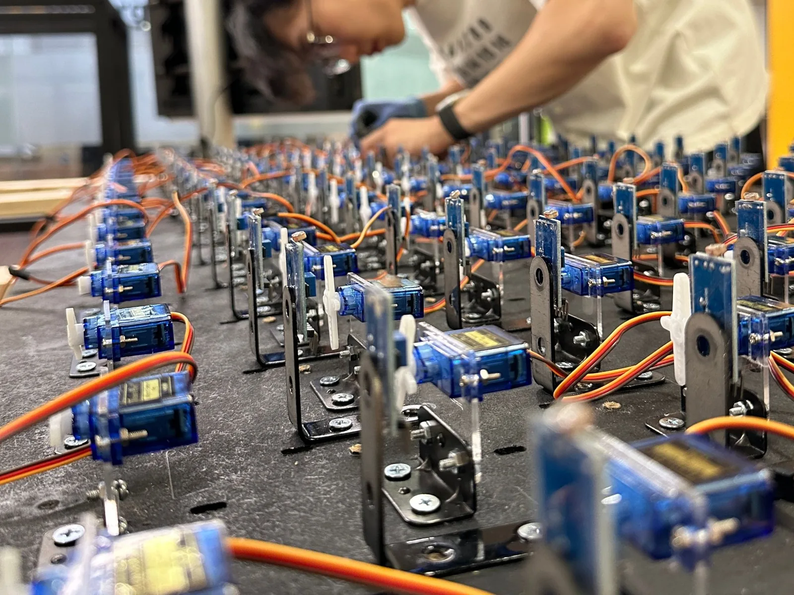 Servos, brackets, and sensors standing in rows on a black board, with a team member assembling in the background.
